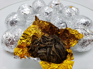 A set of tea balls wrapped in silver foil with one ball partially unwrapped, revealing the dark, compressed tea inside. The foils are placed on a white surface.