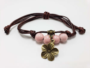 Handmade adjustable cord bracelet with pink ceramic beads and antique brass flower charm on white background