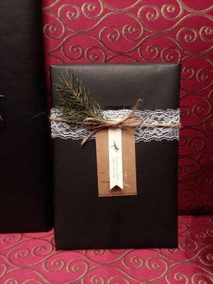 Black gift box wrapped with lace and twine, accented with a sprig of greenery, against a red patterned background. 